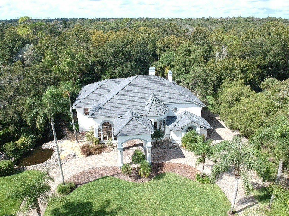 Aerial view of a large, white, two-story house with a gray tile roof, surrounded by trees, landscaped gardens, a pond, and a stone driveway.