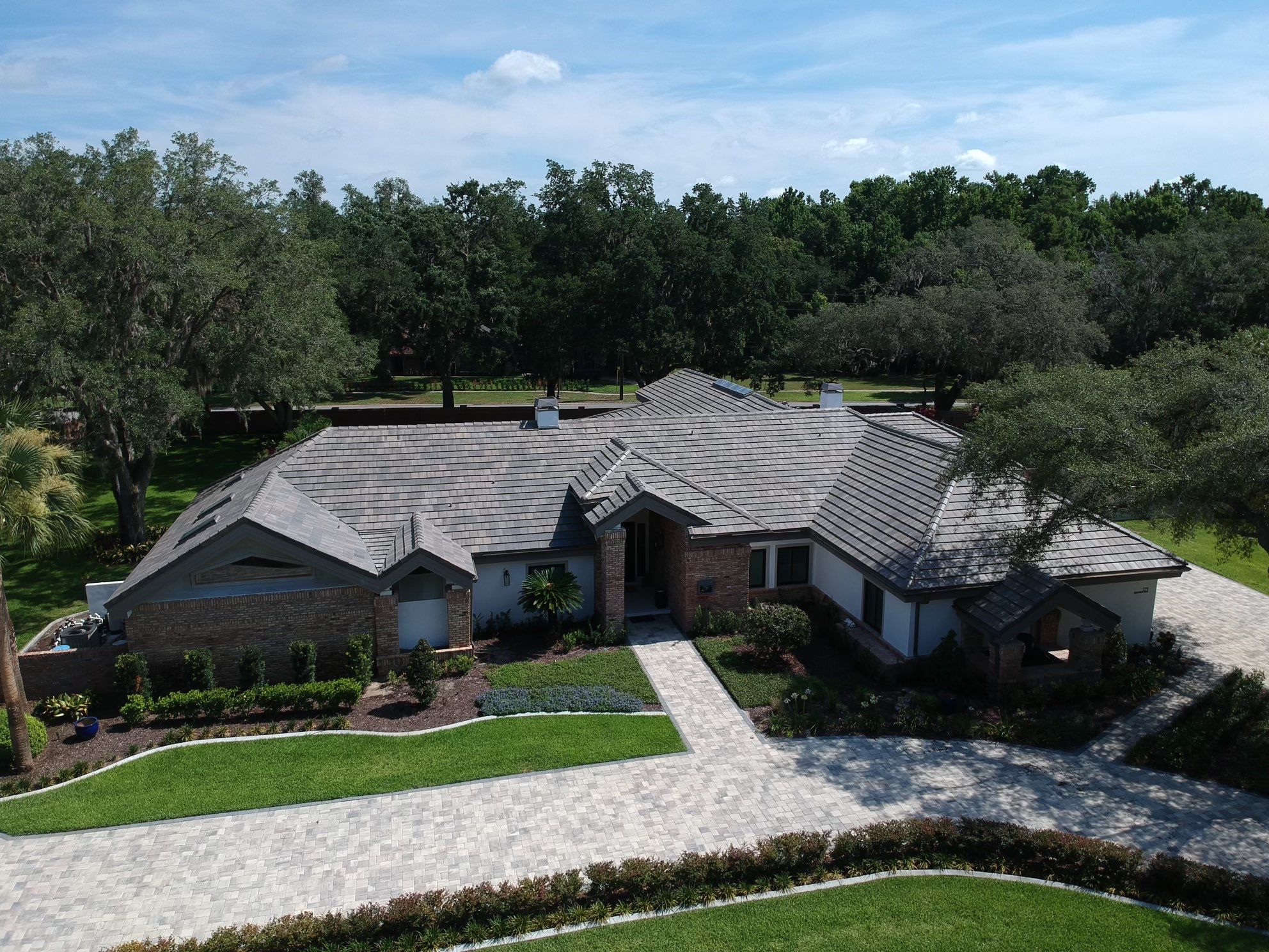 A modern single-story house with a gray tiled roof, brick and stucco exterior, surrounded by landscaped lawns and trees under a blue sky.