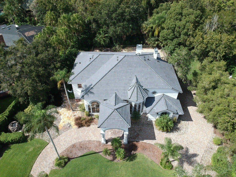 Aerial view of a large two-story house with a tiled gray roof, surrounded by trees, a paved driveway, and landscaped greenery.